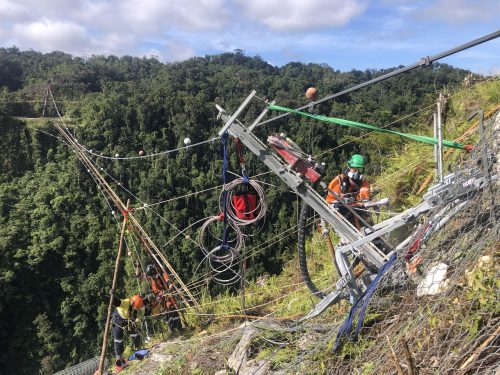Rope access technicians operate heavy machinery on a steep incline for bridge construction, with complex rigging and a dense forest under a bright sky in the background.
