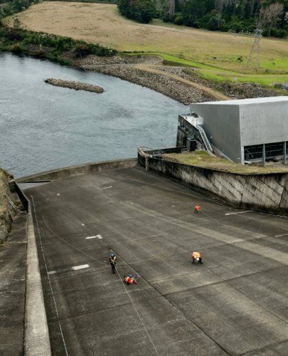 Four rope access technicians are spaced out along the vast concrete spillway of a hydroelectric dam. The grey spillway contrasts with the verdant riverbanks and pastoral landscape. The dam structure is visible to the right, with calm river waters on the left.