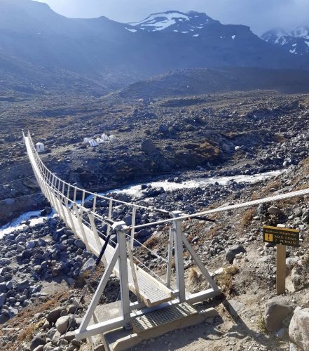 A sturdy suspension bridge spans a rocky, barren landscape with a shallow river running beneath. The bridge, supported by solid metal towers, is marked by a sign indicating a maximum load of one person. In the background, majestic snow-capped mountains loom over the rugged terrain, enhancing the scene's remote and wild atmosphere.
