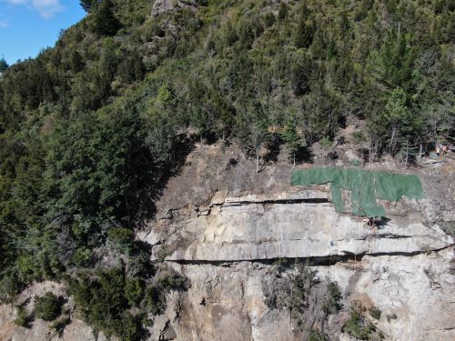 An aerial view showing a cliff stabilisation worksite, with technicians abseiling down the rockface, partially covered with green protective mesh, amid lush green forest.