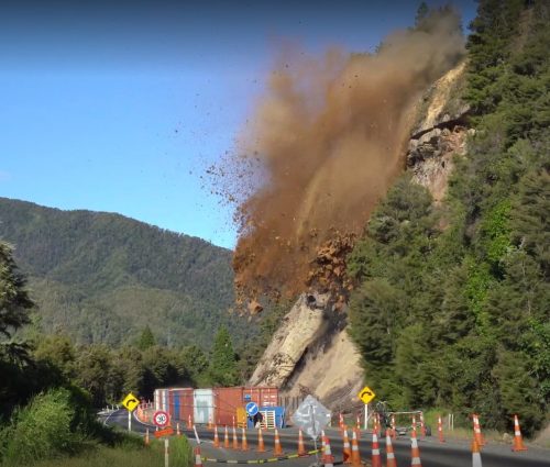 Controlled rock blasting is in progress on a hillside next to a road, with a dust cloud rising above traffic cones and safety barriers set against a backdrop of dense forest.
