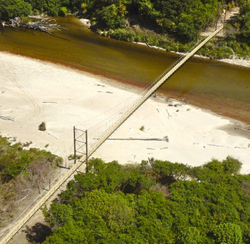 Aerial view of a narrow suspension bridge spanning a river and adjacent sandy beach area, surrounded by dense foliage. The bridge features a metal frame and wooden planks, connecting two lush green forested areas, with the calm river reflecting the bright sunlight.