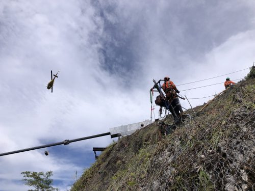 Rope access workers on a steep slope with construction equipment as a helicopter hovers above against a cloudy sky, showcasing precise coordination in a challenging environment.