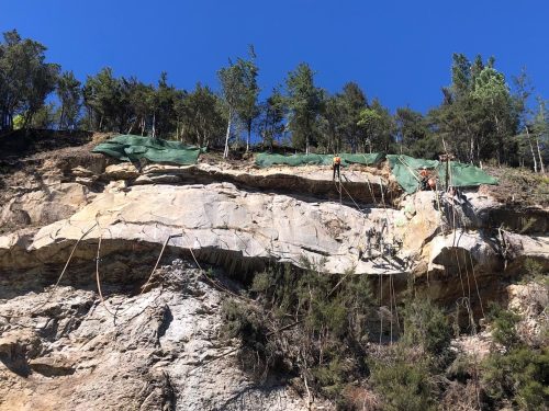 Rope-access technicians secure a rocky cliff face with green mesh to prevent rockfall under a clear blue sky.