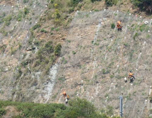 Technicians in orange overalls wearing harnesses carefully navigate a sparse, sloping terrain with geotextile covering dotted with soil nails and anchor plates. They are attached to yellow safety ropes, highlighting the precision required in their movements while conducting geotechnical stabilisation to protect the landscape from erosion.