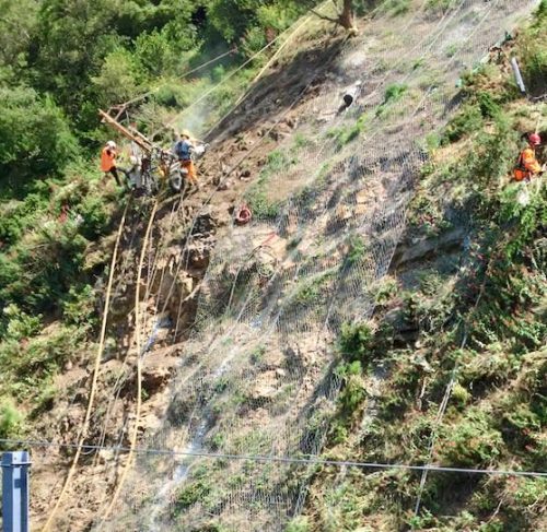 An aerial view of a steep slope where a team of workers in safety gear is installing rockfall mesh. Amidst scattered vegetation, the slope reveals the ongoing construction with a crisscross of ropes and partially laid mesh, symbolising the intricate work of ensuring slope stability and preventing soil erosion in a natural setting.