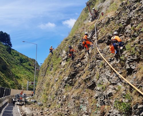 A team of rope access technicians clad in orange and white safety gear methodically work on a steep, rocky embankment. They are secured by harnesses as safety measures are installed along the hillside above a highway. Their skilled coordination is evident as they work at various heights, with ropes and equipment laid out, demonstrating their commitment to maintaining and securing challenging terrains.