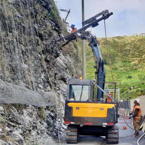 A rockfall protection operation with a worker operating a hydraulic boom from atop a yellow and black crane. The crane extends towards a steep, rocky hillside to install wire mesh for slope stabilisation. Another worker on the ground, in protective gear, oversees the process. Rolls of mesh await deployment, illustrating the proactive measures taken to safeguard against rockslides.