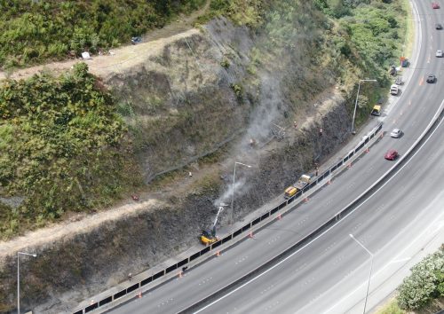 An aerial view of a highway adjacent to a steep hillside where rockfall mitigation work is underway. Dust rises from the area where workers in bright safety gear use machinery to clear and stabilise the slope. Traffic cones partition the work zone from the active lanes, where vehicles continue to pass, highlighting the careful balance between infrastructure maintenance and traffic flow.