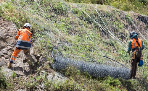 Two professionals in high-visibility gear and helmets work on a steep slope installing rockfall netting. The metallic mesh is draped over the terrain and anchored by steel cables to protect against debris fall. This intervention highlights safety measures undertaken in vulnerable areas, with the workers carefully navigating the rugged landscape to secure the netting.