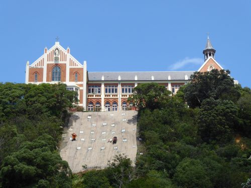 A captivating image showcasing workers scaling a reinforced geotextile-covered slope beneath an impressive red and cream church with a statue and a steeple. The hillside installation highlights the melding of technology and nature, with dense greenery on either side. This setting exemplifies modern engineering integration into the historical and natural landscape.