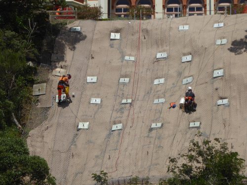 Technicians in high-visibility gear work meticulously on a hillside with a large geotextile mesh anchored by white plates. The workers employ specialised equipment for the installation, which is visible by their careful positioning on the steep terrain. In the background, the upper portion of a historical building adds a cultural element to the scene of rigorous environmental engineering.