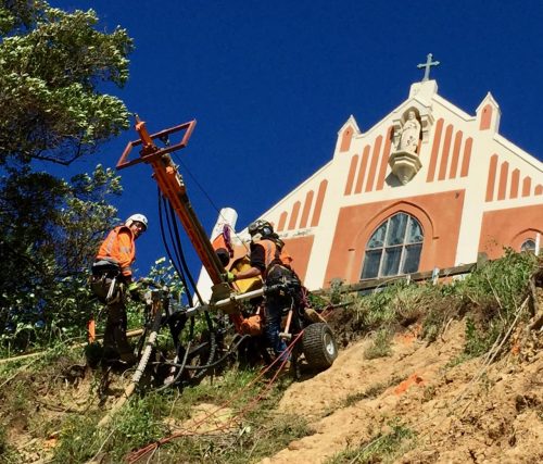 Technicians in high-visibility clothing operate soil drilling equipment on a slope before a historic red and cream church. The clear blue sky provides a vivid backdrop for the workers who are focused on stabilising the ground. This image illustrates the careful balance between preserving cultural architecture and undertaking essential geotechnical work.
