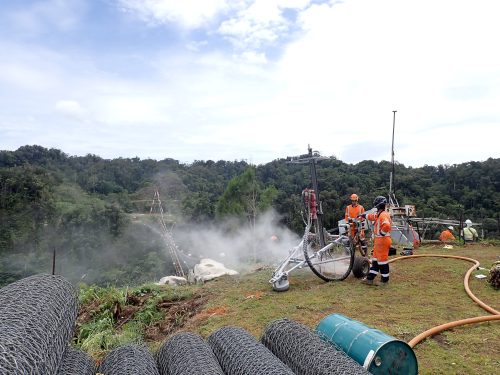 Rope access workers and engineers with machinery at a bridge construction site, with rolls of cable in the foreground and a forested valley beyond under a cloudy sky.