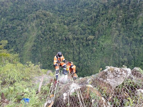 Rope access technicians installing rockfall netting on a steep slope with a dense forest backdrop.