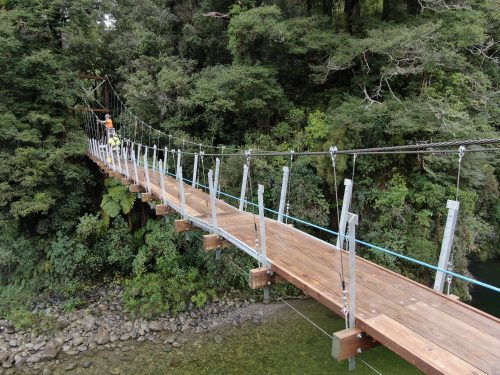 A suspension bridge with wooden planks and steel cables, with workers conducting inspections, stretches across a river bordered by a dense forest.