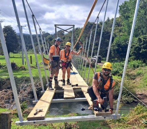 Three technicians in high-visibility gear smiling on a partially reconstructed suspension bridge, with one kneeling on the wooden walkway, surrounded by lush countryside.