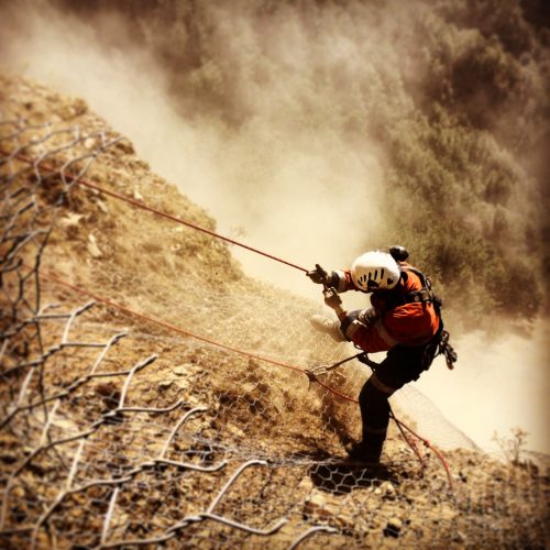 A rope access technician is descending a steep, dusty slope anchored by ropes to a rockfall protection mesh. The worker wears a white helmet, an orange high-visibility jacket, and a harness. A cloud of dust envelopes the area, hinting at the challenging conditions of the environment.