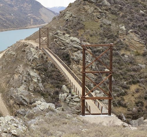 A rustic suspension bridge with a wooden walkway traversing a rugged landscape, with a backdrop of a serene lake and mountainous terrain.