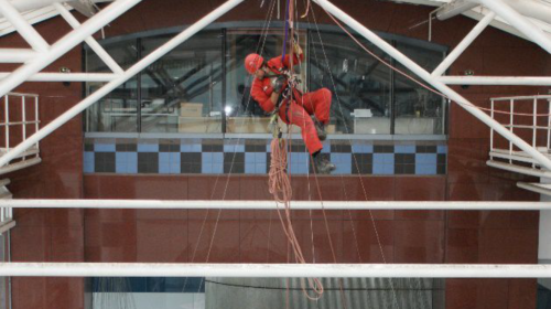 Suspended mid-air within a geometric steel structure, a rope access technician in red coveralls conducts maintenance. The professional is secured by a complex system of ropes, with the indoor environment visible through the glass facade in the background, highlighting the blend of architecture and specialised high-altitude work.