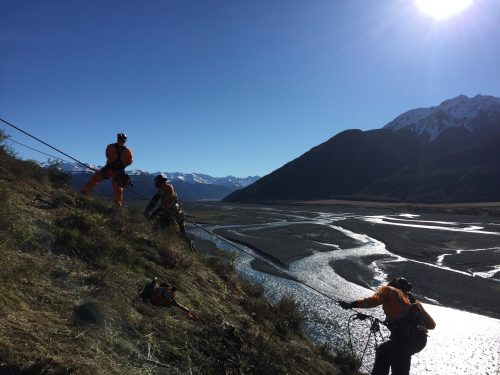 Rope access technicians in bright orange safety gear are secured with harnesses on a steep grassy slope. They are working above a winding river valley with a backdrop of majestic snow-capped mountains under a clear blue sky, with the sun casting a brilliant glare on the water.