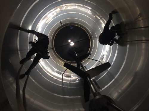 Two technicians with safety harnesses inspect a large, cylindrical metal shaft silhouetted against a luminescent circular opening at the top. Their ropes and equipment cast long shadows on the curved reflective interior walls, conveying depth and scale.