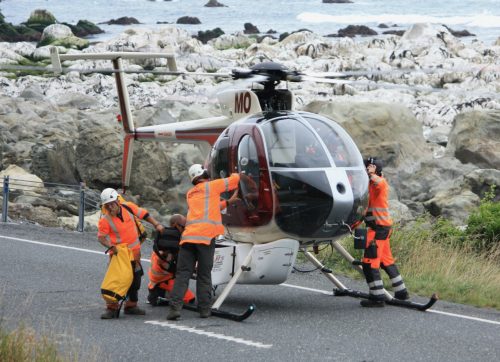 Rope access technicians in high-visibility uniforms conduct operations with a small red and white helicopter parked on a coastal road. The rocky shoreline and ocean waves provide a rugged backdrop to this precise and carefully coordinated activity.