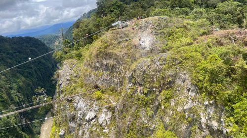 An expansive view of a rockfall protection operation on a cliff, with cables strung across the scene and workers on a craggy slope amidst lush vegetation with a valley stretching into the distance.